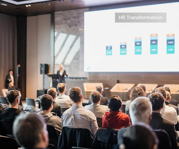 Audience attending a business presentation with charts and an “HR Transformation” overlay.