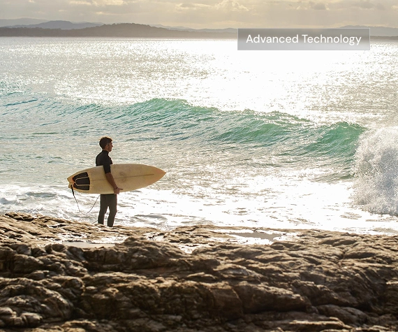 Surfer holding a board on a rocky shore beside ocean waves with an “Advanced Technology” overlay.